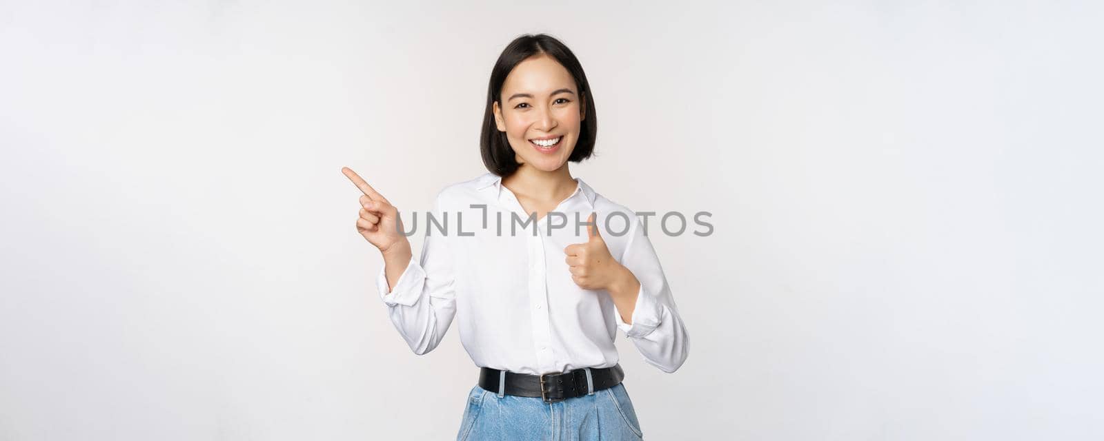 Image of young asian business woman, smiling while pointing finger left and showing thumbs up, recommending product, praise, standing over white background by Benzoix