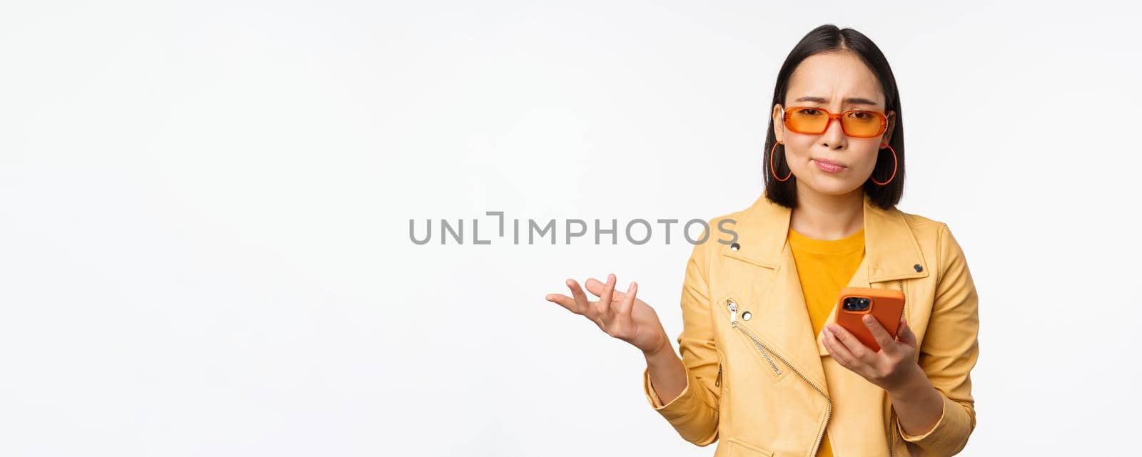 Portrait of confused asian girl in sunglasses, holding smartphone, using mobile phone and looking puzzled, standing clueless over white background by Benzoix