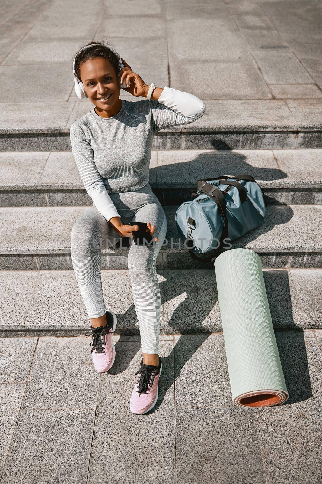 Sportswoman with sportive bag and yoga mat smiling at camera, resting outdoors