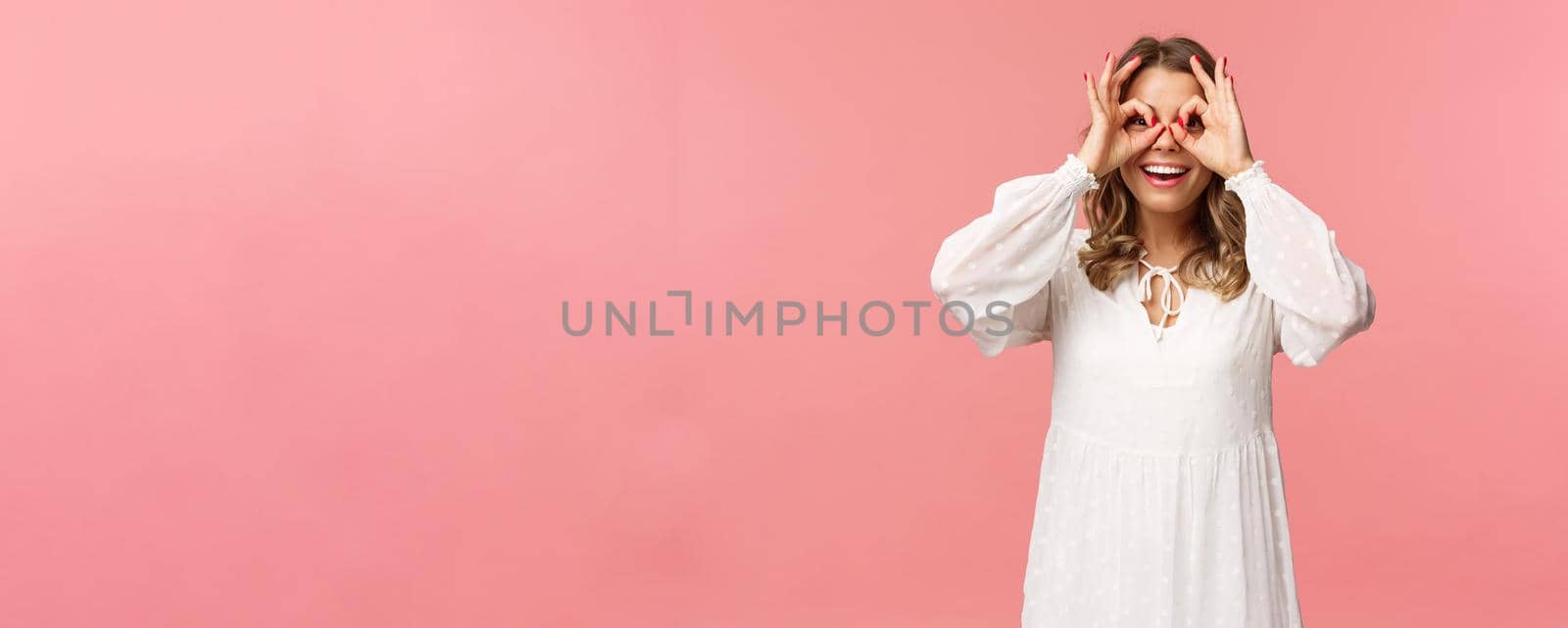 Portrait of dreamy cute and funny young blond girl seeing something interesting, looking from okay signs as making glass-mask with fingers over eyes, smiling amused, pink background.