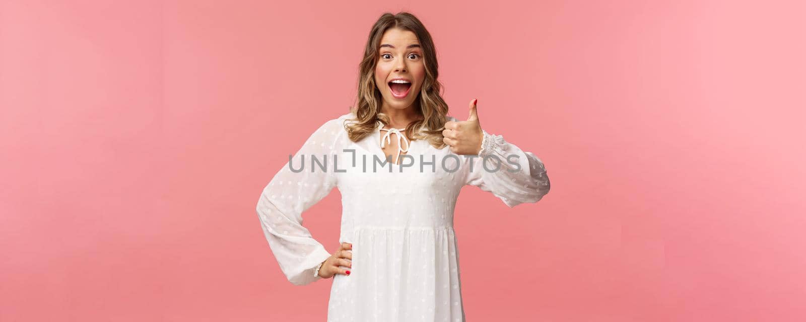 Great idea. Upbeat, enthusiastic blond caucasian girl in white spring dress, hear good plan, show thumbs-up and nod in approval, agree and recommend, satisfied with choice, stand pink background by Benzoix