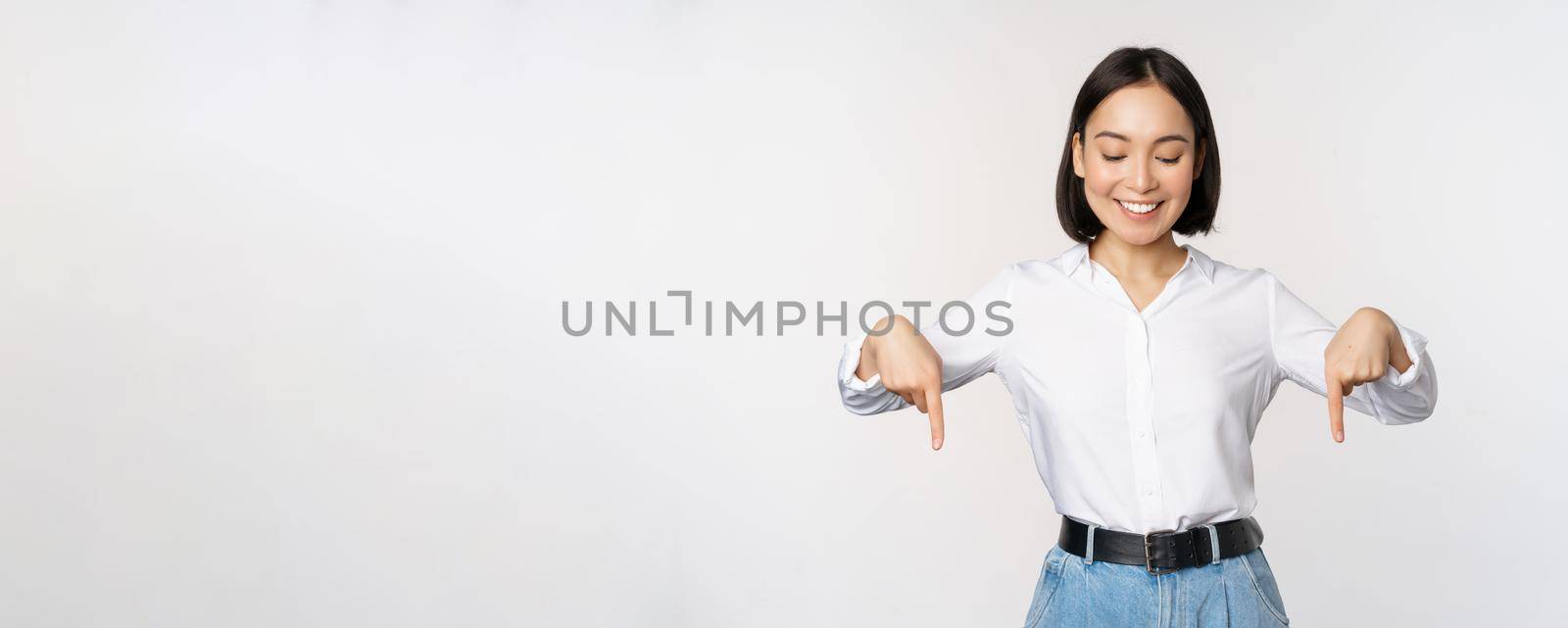Portrait of happy asian woman pointing fingers down and looking below at advertisement, showing info banner, advertising, standing over white background.