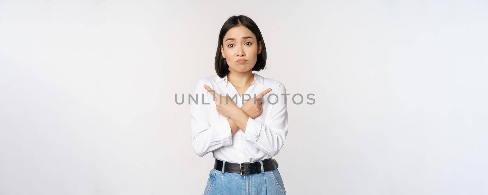 Confused sad asian girl cant decide, pointing fingers left and right, sulking and shrugging indecisive, cannot choose, standing over white background by Benzoix
