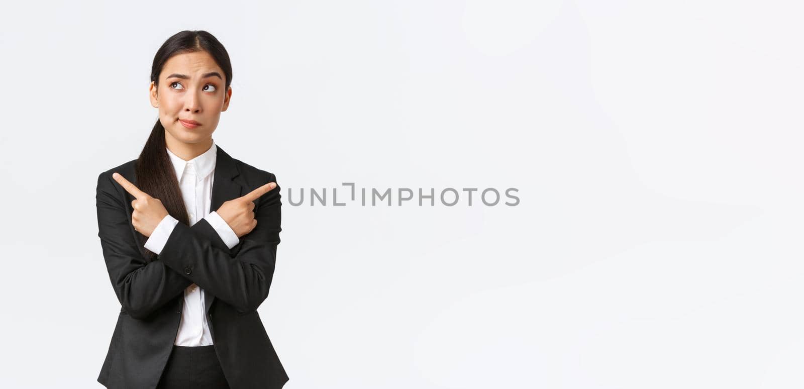 Skeptical and doubtful asian businesswoman hesitating making decision, smirk uncertain looking upper left corner while pointing fingers sideways, looking confused and unsure, white background.