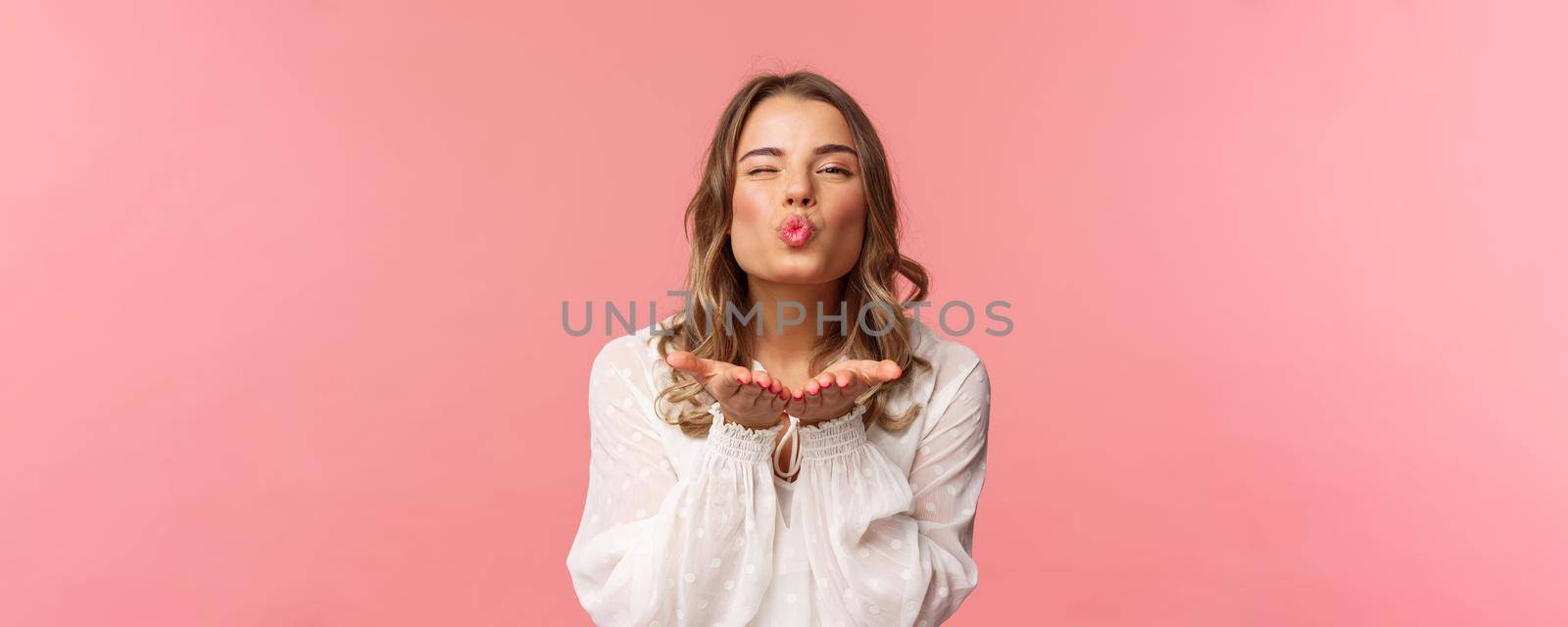 Close-up portrait of lovely feminine girlfriend enjoying spring fine day, sending air kiss at camera, winking and folding lips in mwah, hold hands near mouth, pink background.