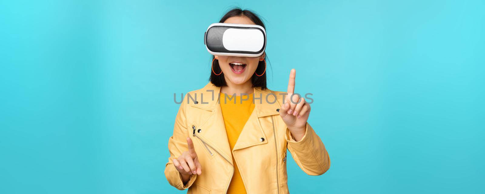 Young asian woman using virtual reality glasses, using VR headset, standing amused against blue background. Copy space