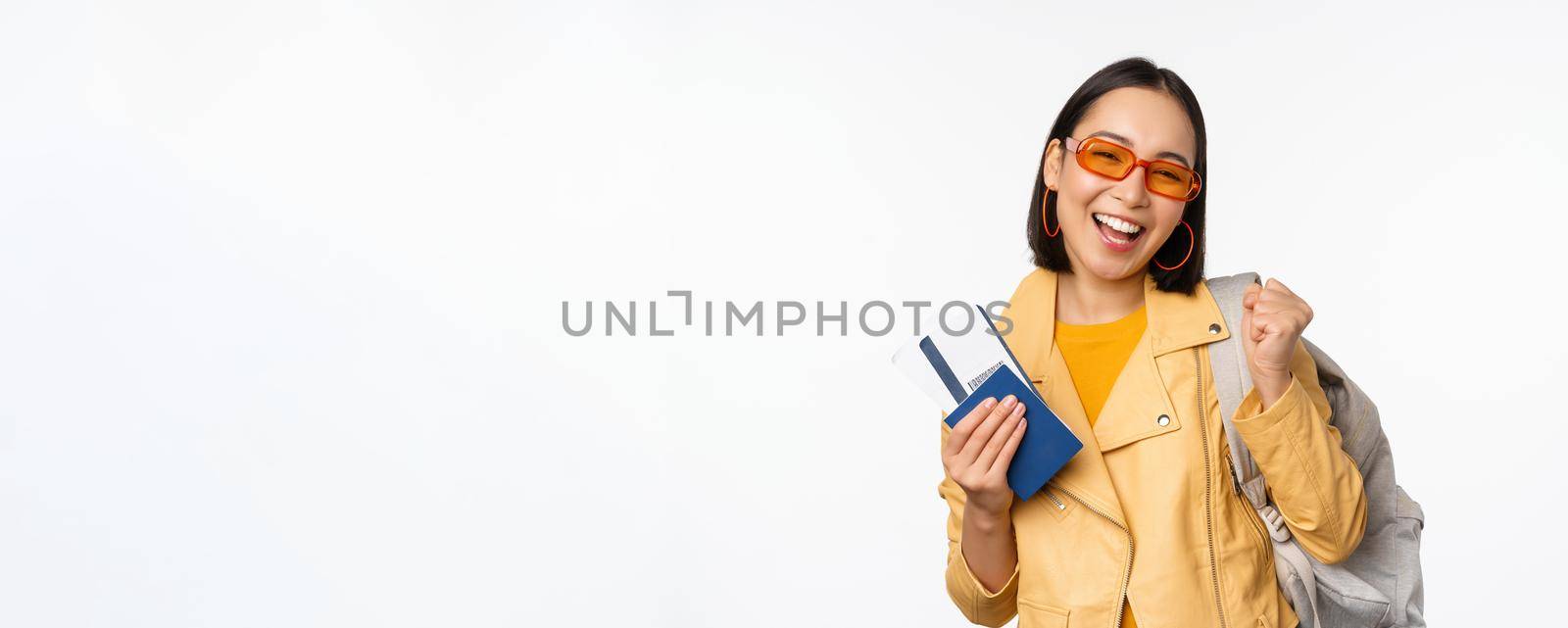 Happy asian girl going on vacation, holding passport and flight tickets, backpack on shoulder. Young woman tourist travelling abroad, standing over white background.