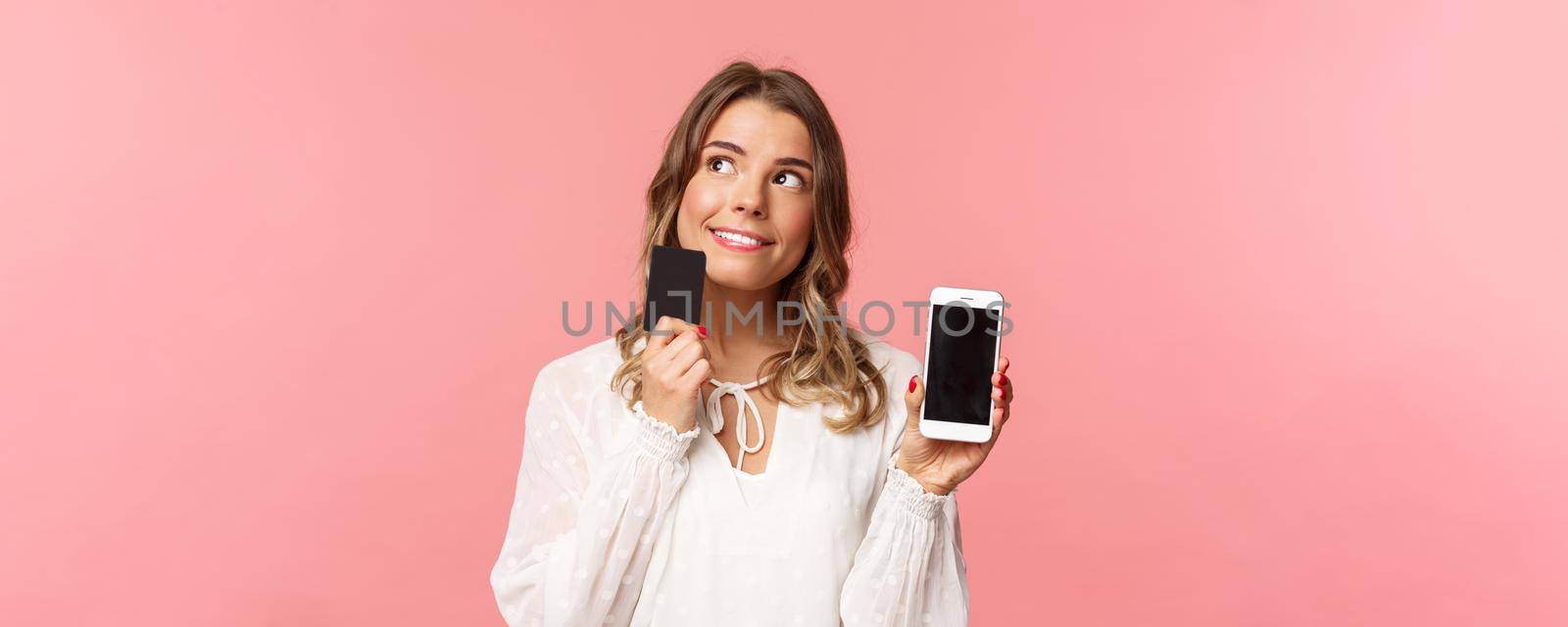 Finance, shopping and technology concept. Close-up portrait of thoughtful, dreamy attractive blond girl in white dress, thinking what buy, order something online shop, hold smartphone and credit card.