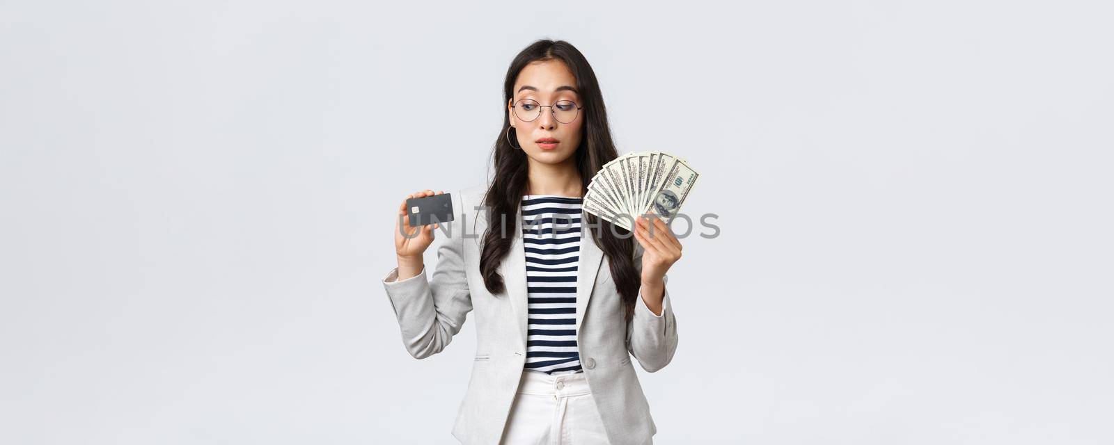 Business, finance and employment, entrepreneur and money concept. Indecisive cute asian office lady thinking about putting cash on deposit, looking curious at credit card, white background.