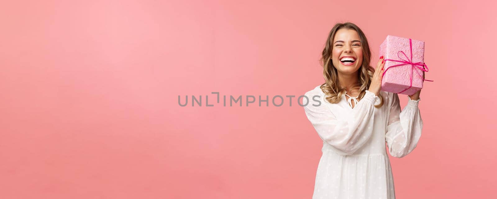 Holidays, celebration and women concept. Portrait of happy charismatic blond girl shaking gift box wondering whats inside as celebrating birthday, receive b-day presents, pink background by Benzoix