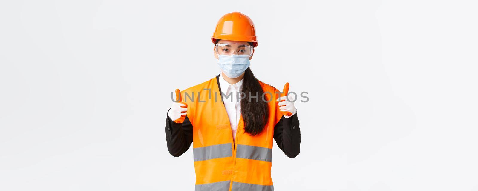 Covid-19 safety protocol at enterpise, construction and preventing virus concept. Confident female asian industrial worker, engineer in face mask and helmet showing thumbs-up, all good.