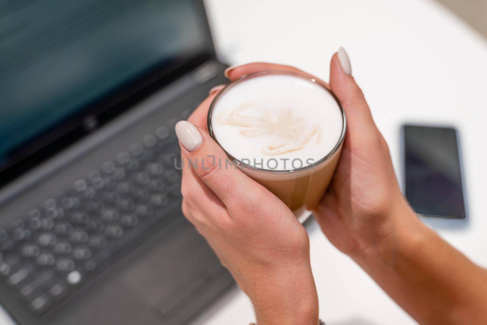 Close-up of beautiful female hands holding a large white cup of cappuccino. A business woman sits in a cafe working at a computer