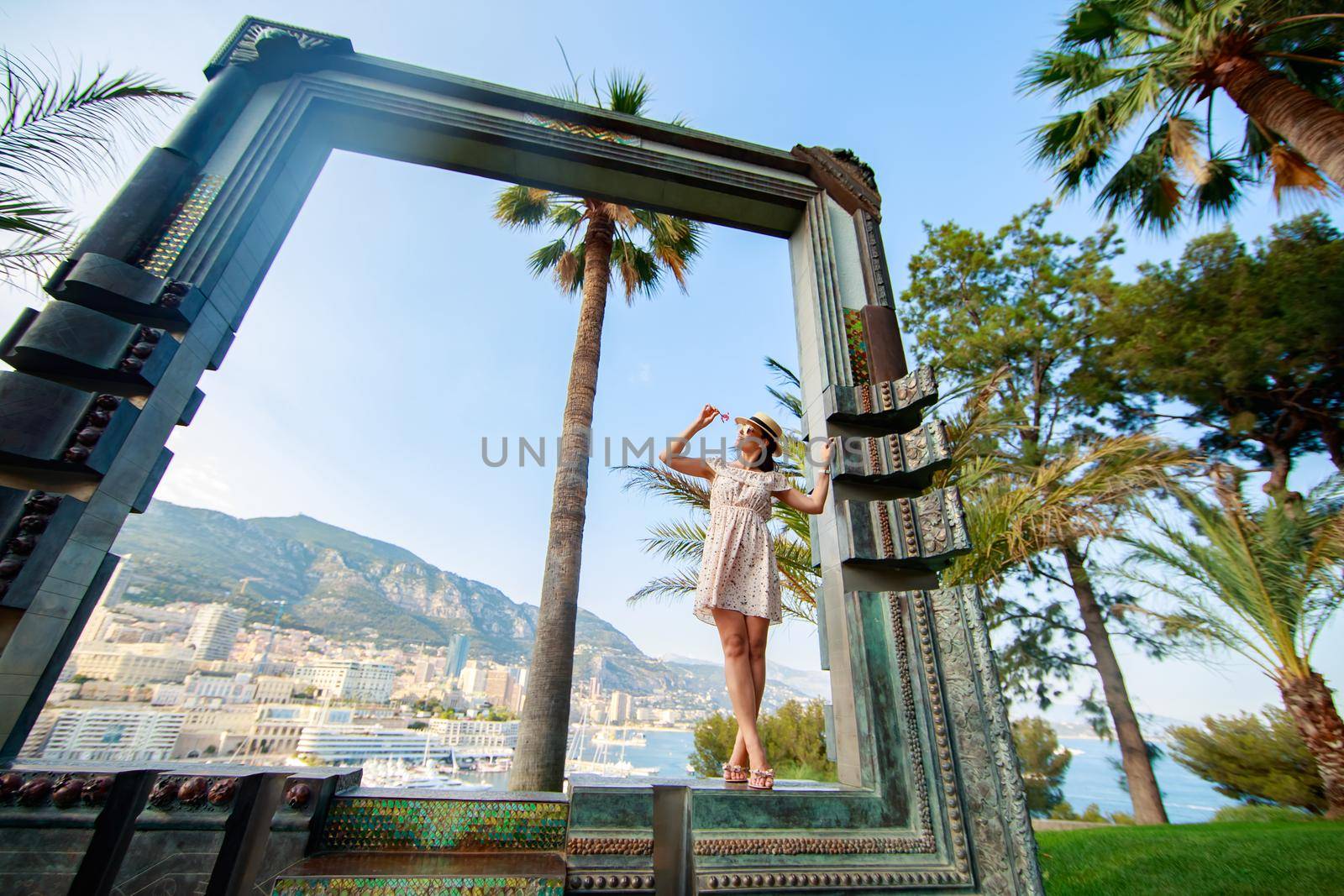 a bright beautiful girl in a light dress and hat walks near landmark of Monaco in sunny weather, she smells a flower, a residential area on background. High quality photo