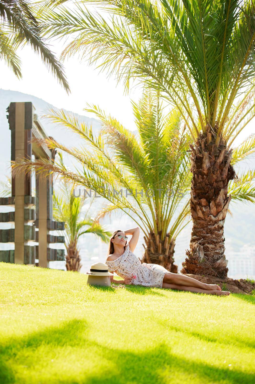 a bright beautiful girl in a light dress and hat lies on the grass under a palm tree in Monaco in sunny weather in summer, streets of old town of Monaco. High quality photo