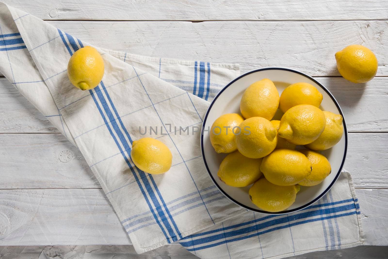 yellow lemons in a plate and on a white towel with blue stripes on white wooden boardsHigh quality photo