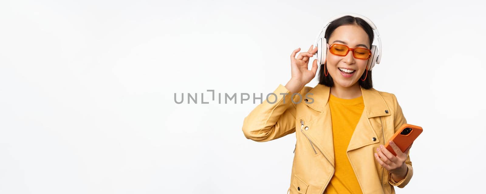 Happy stylish asian woman in sunglasses, listening music in headphones, holding smartphone, singing and dancing, standing over white background.