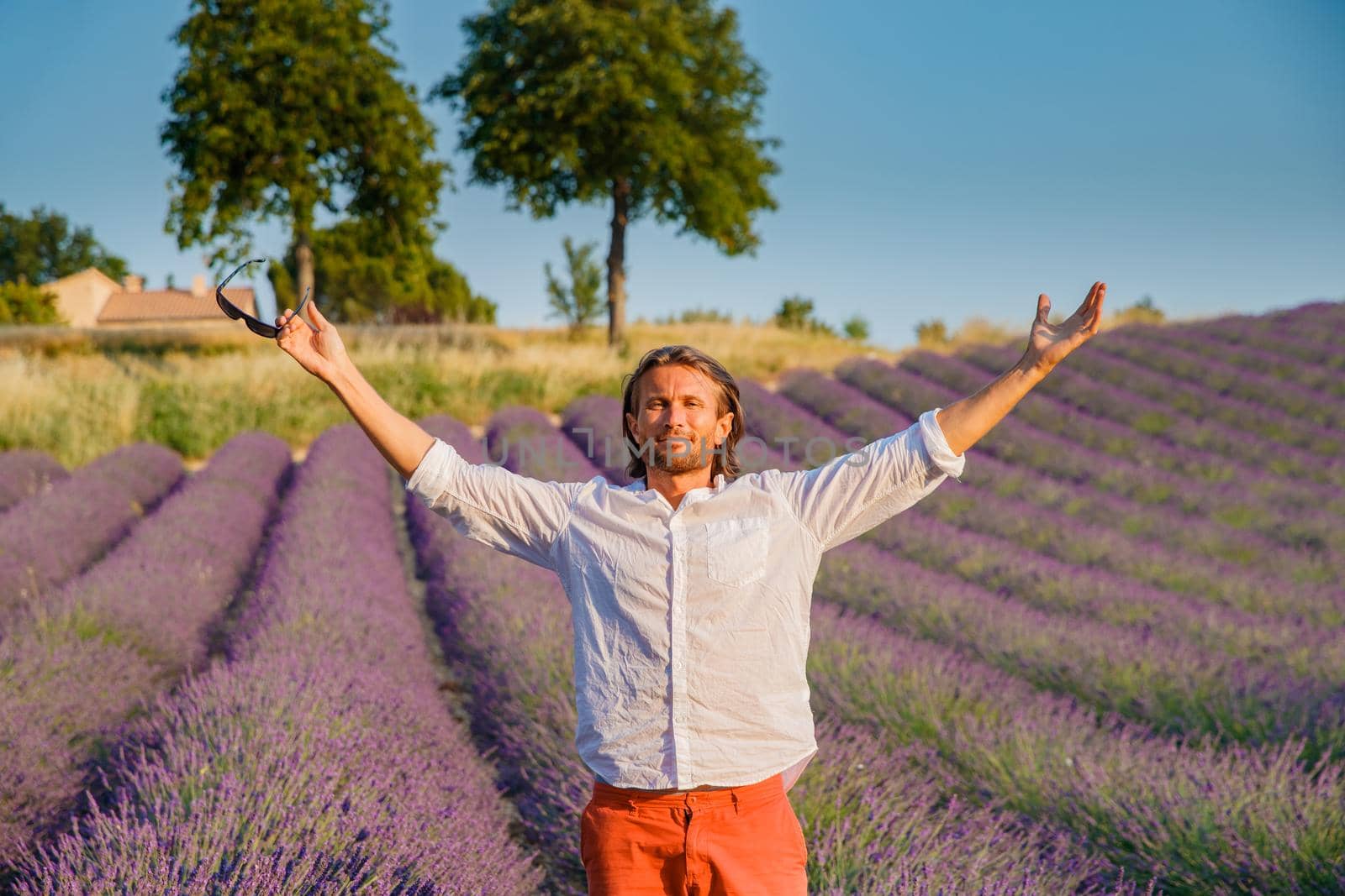 The handsome brutal man with long brunette hair poses in the field of lavender in provence near Valensole, France, clear sunny weather, in a rows of lavender, red shorts, white shirts, blue sky by vladimirdrozdin