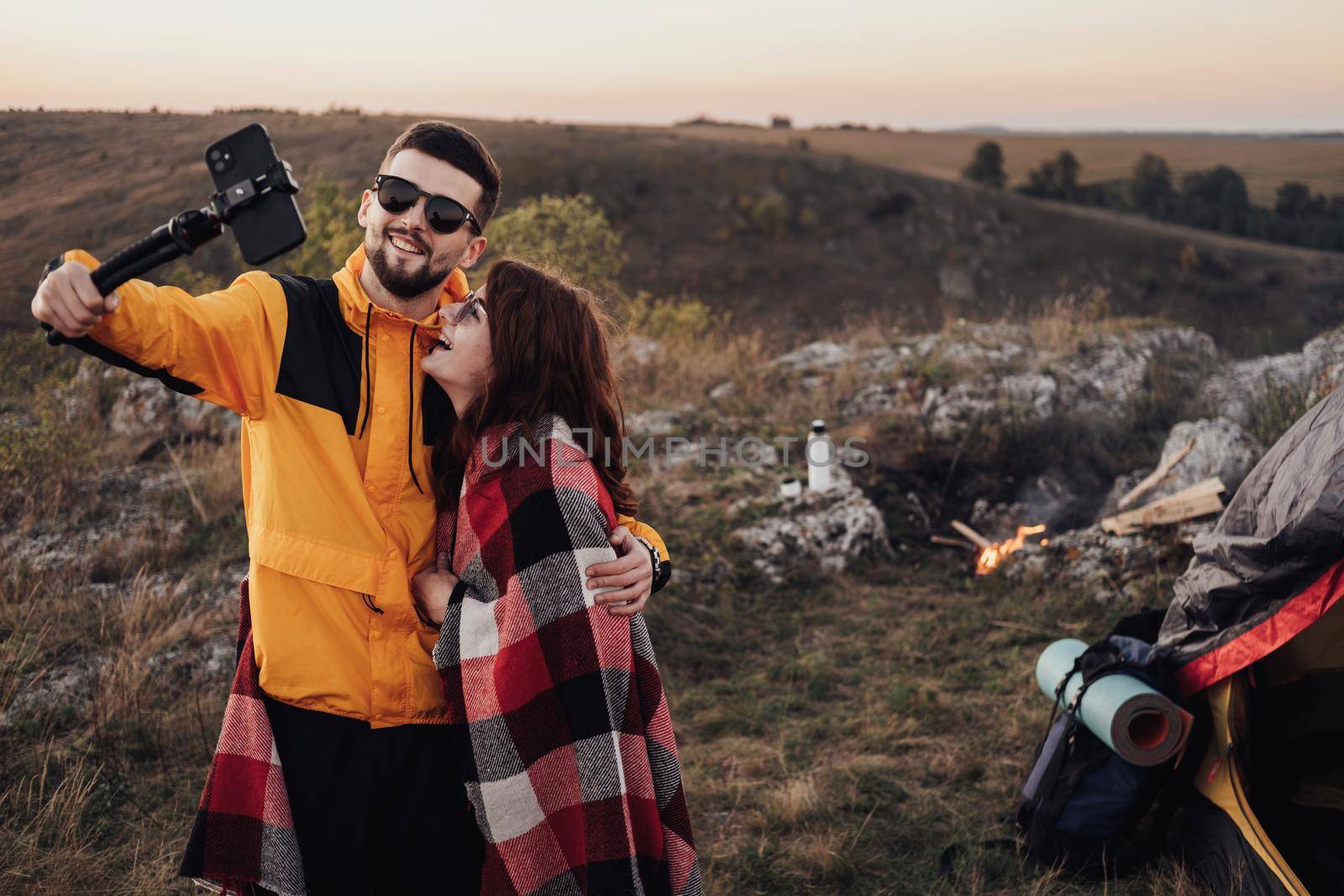 Young Traveler Couple Making Selfie on Top of Hill at Background of Campfire and Tent in Evening