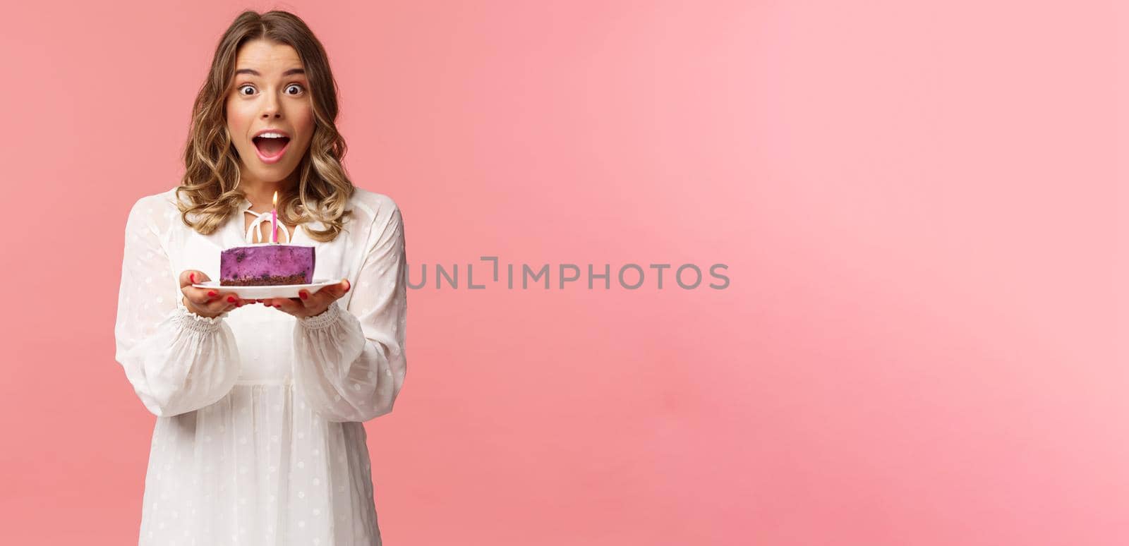 Holidays, spring and party concept. Happy cheerful good-looking blond woman celebrating birthday, holding piece cake with lit candle, making wish, look amused standing pink background by Benzoix