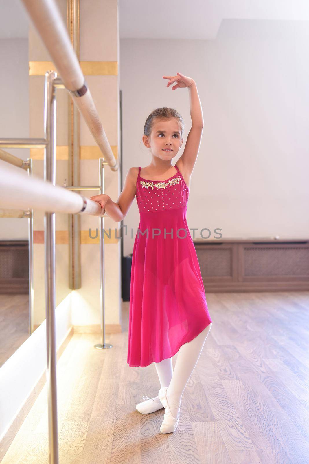 charming little girl dreams of becoming a ballerina. The girl in the pink dress is dancing, holding on to the bar.Baby girl is studying ballet. by Nickstock