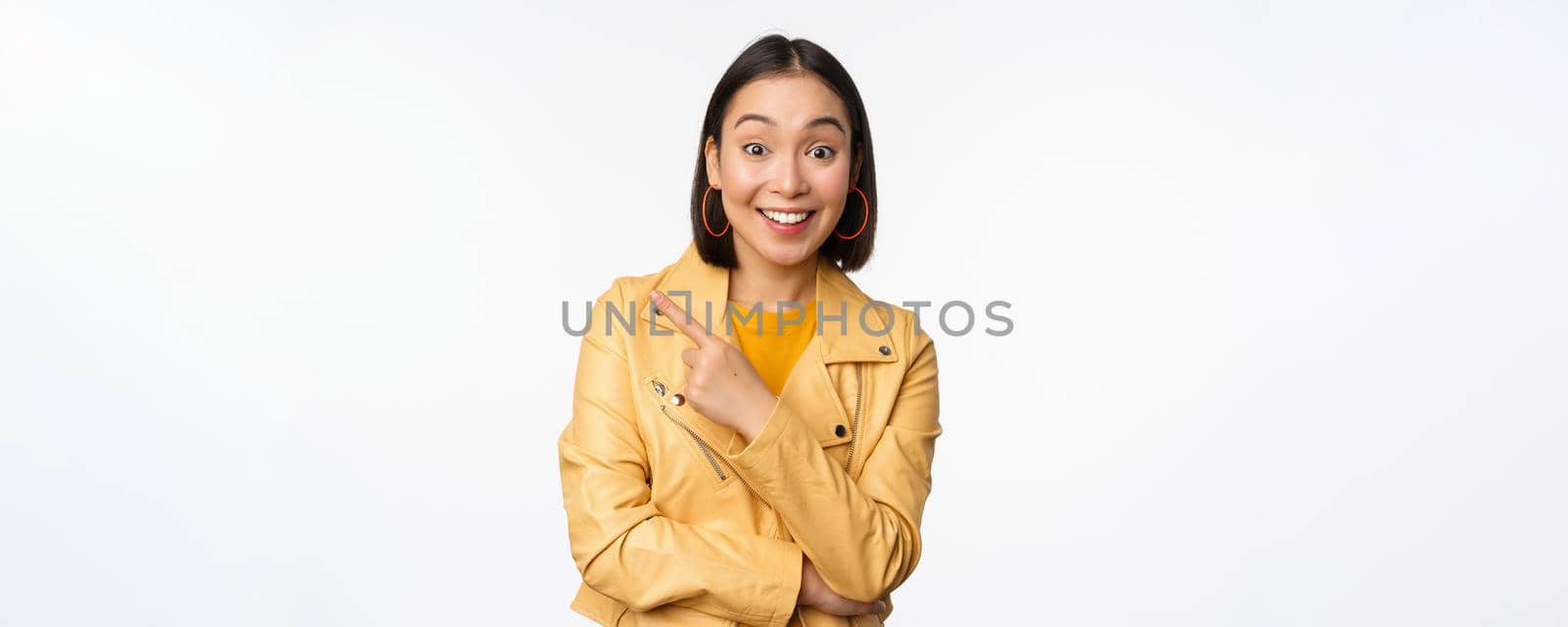 Portrait of beautiful asian woman express intereest and excitement about advertisement, store discount, pointing finger left, looking amazed, standing over white background.