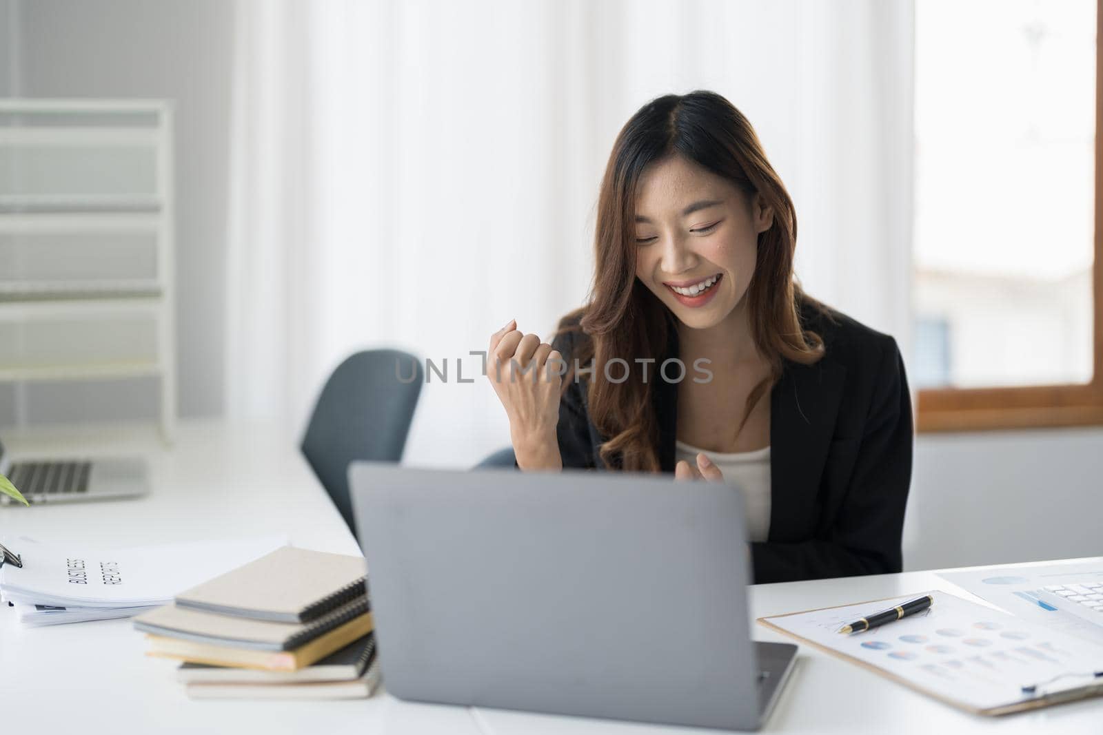 Happy young asian business woman in suit looking at laptop excited by good news online, lucky successful winner man sitting at office desk raising hand in yes gesture celebrating business success win result.