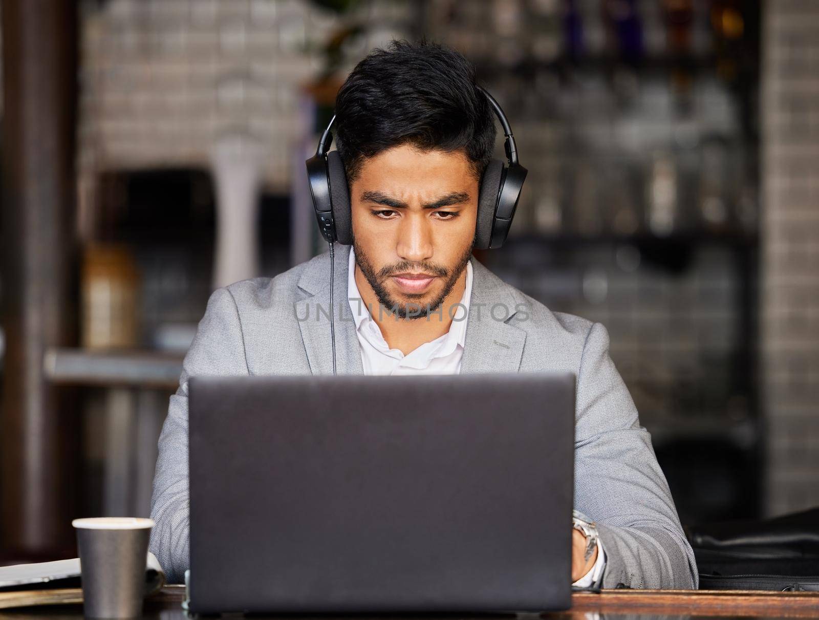 Shot of a young businessman wearing headphones while using a laptop at a cafe.