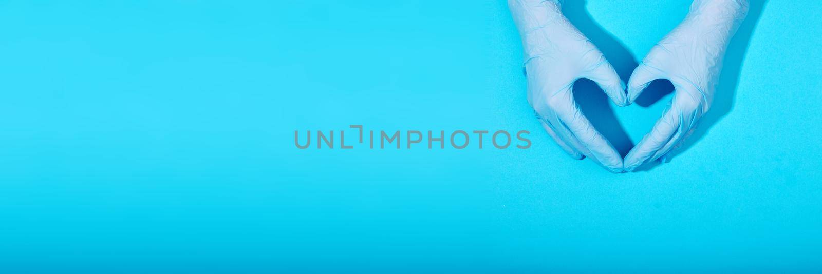 Hands in latex rubber gloves lie in the shape of a heart sign on a blue background with clear shadows. Banner for website with copy space