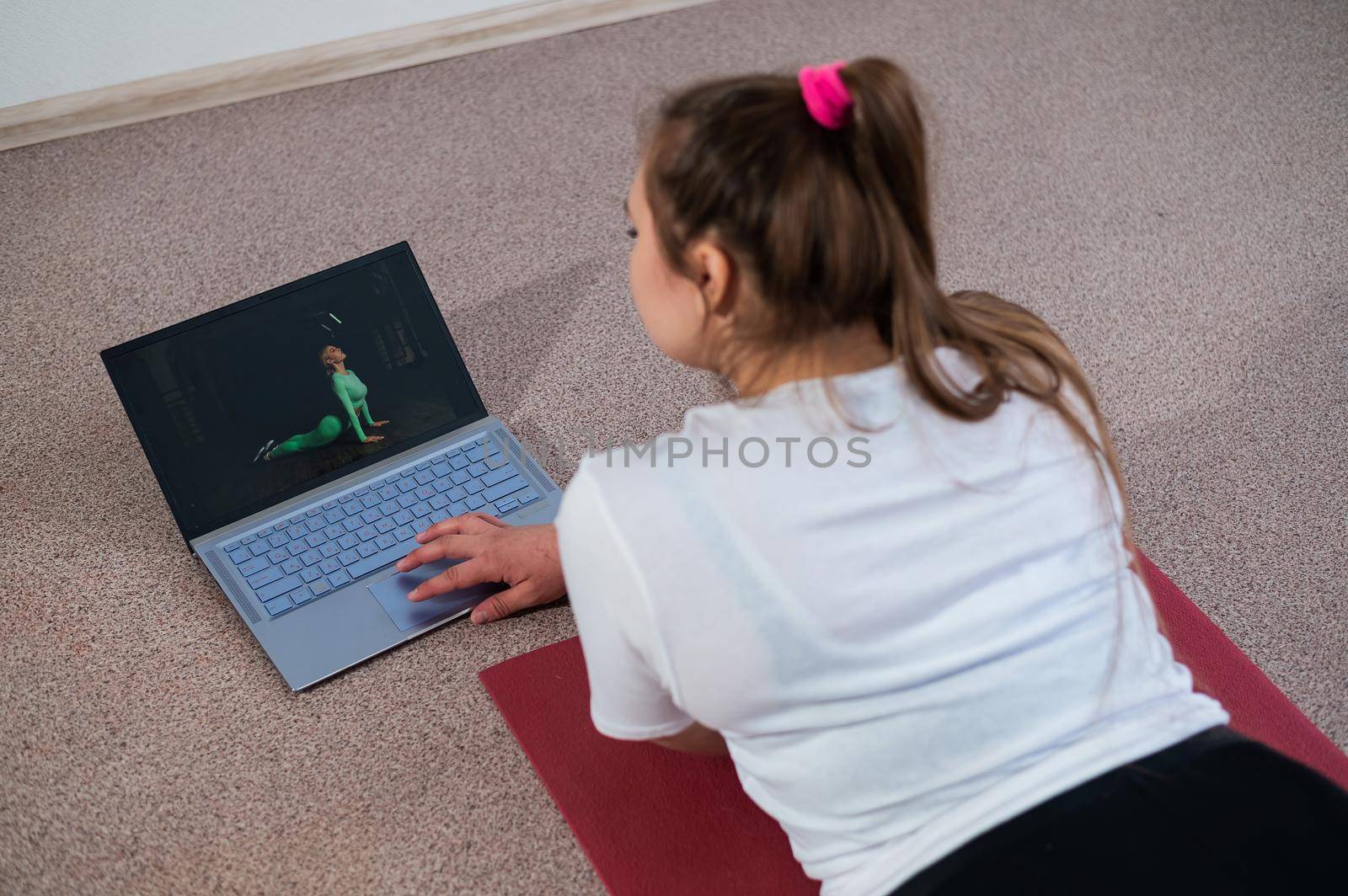A chubby young woman is watching an online fitness lesson on a laptop. Distance sports training