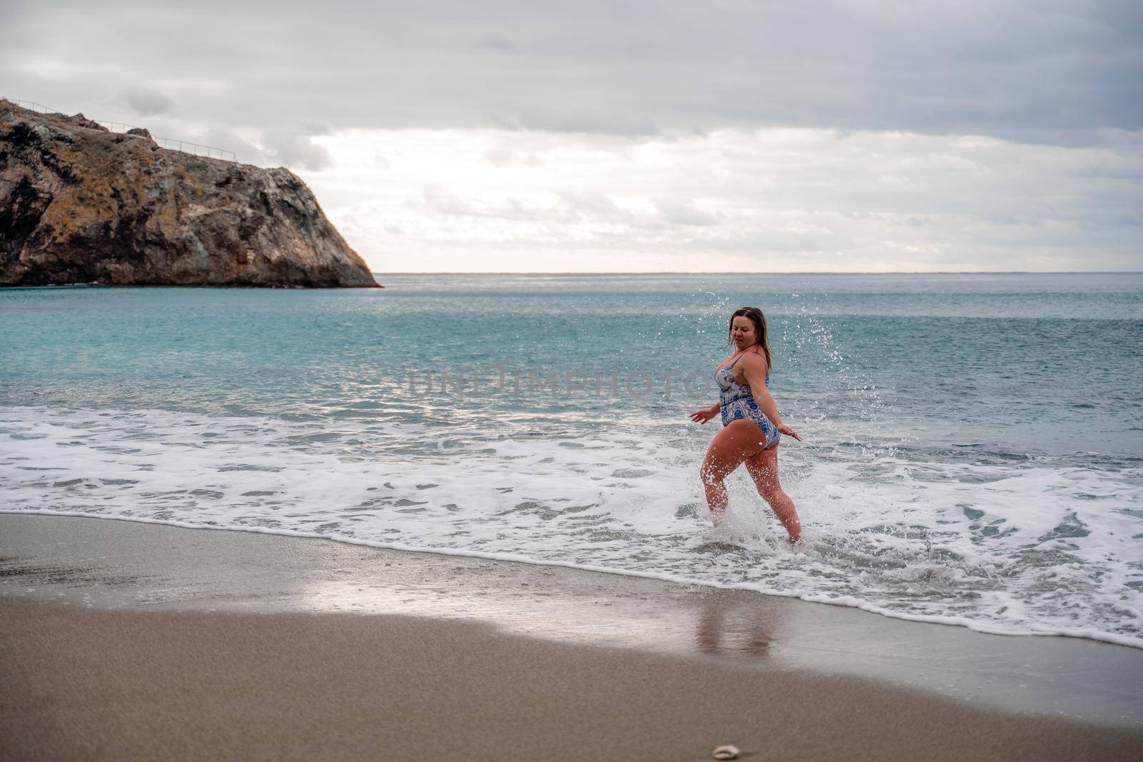 A plump woman in a bathing suit enters the water during the surf. Alone on the beach, Gray sky in the clouds, swimming in winter