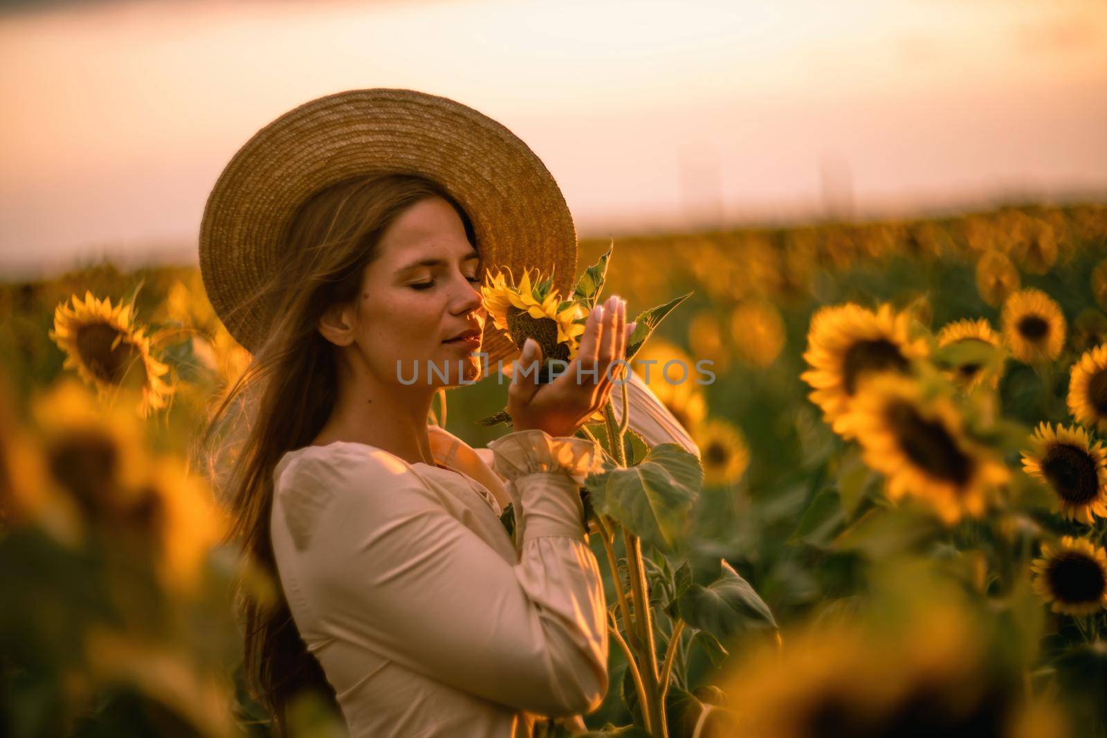 Beautiful middle aged woman looks good in a hat enjoying nature in a field of sunflowers at sunset. Summer. Attractive brunette with long healthy hair. by Matiunina
