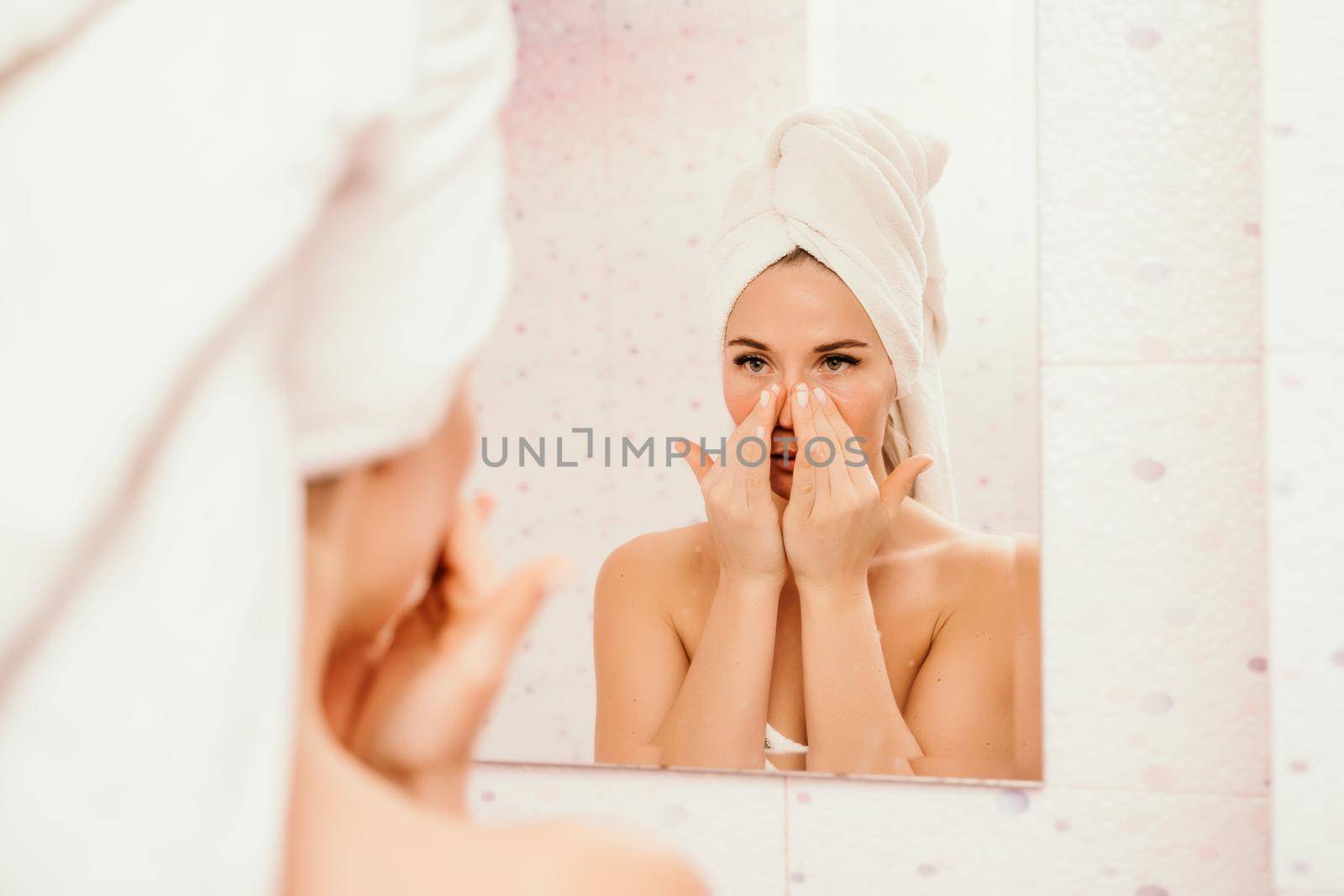 Young beautiful woman using face cream moisturizing lotion after bath. Pretty attractive girl in a towel on her head stands in front of a mirror in a home bathroom. Daily hygiene and skin care.