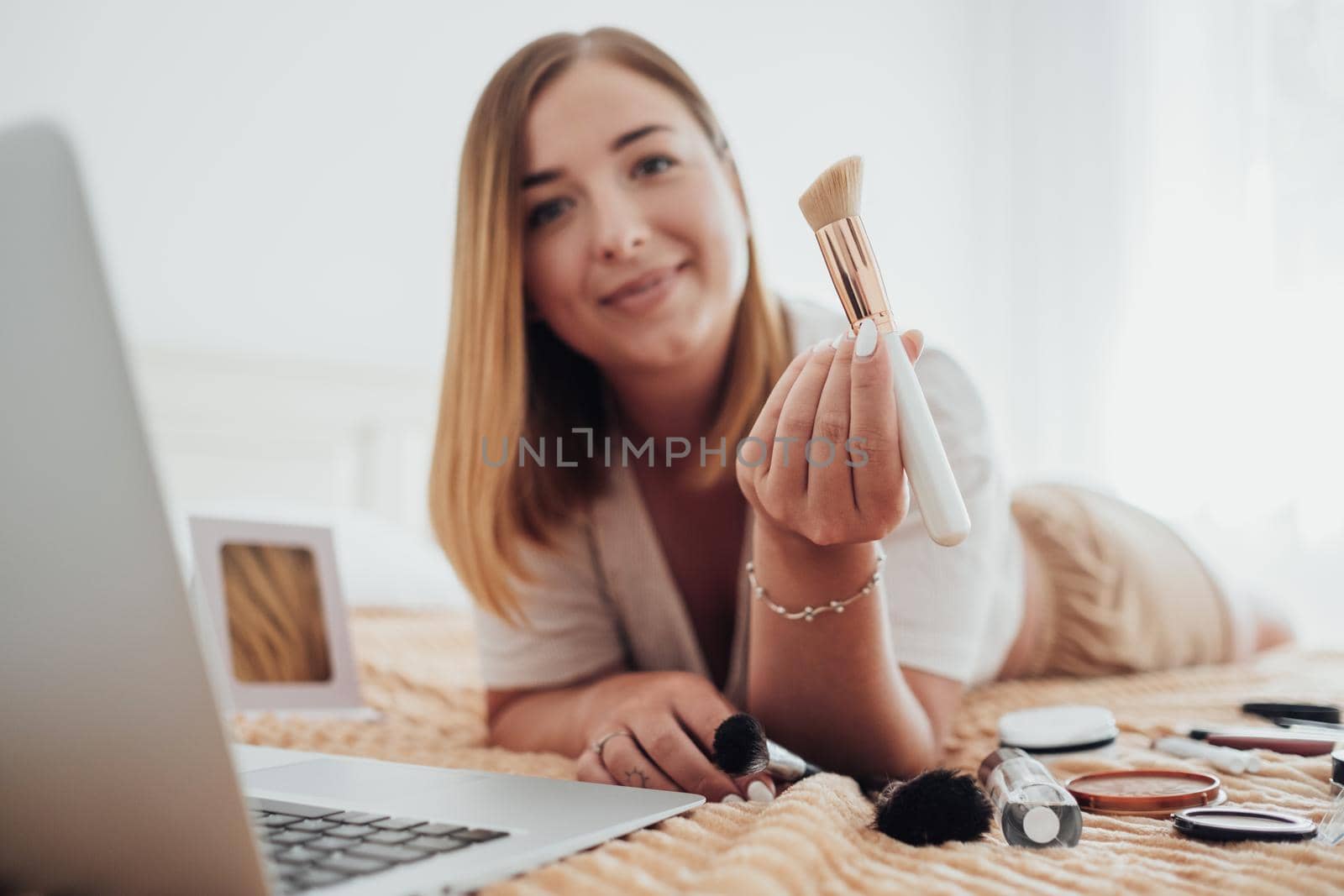 Makeup Brush in Focus, Young Woman Laying on Bed with Cosmetics and Looking Into Camera During Her Online Master Class Through Laptop