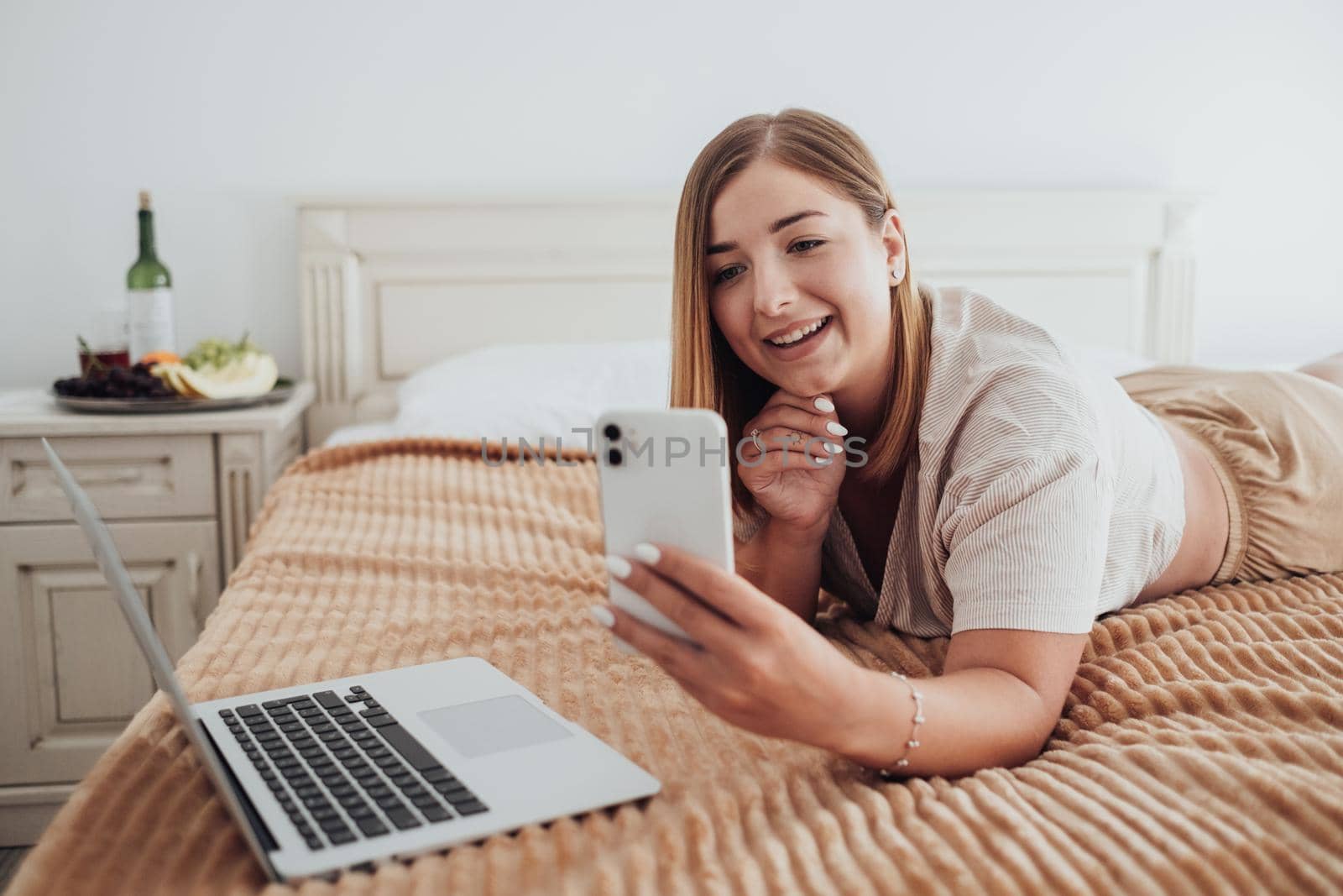 Beautiful Caucasian Woman Laying on Bed in Hotel Room While Using Smartphone and Laptop for Video Call