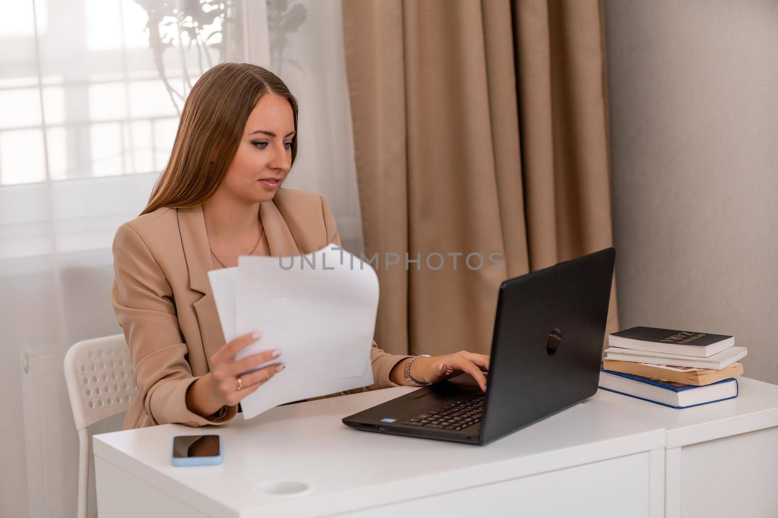 European professional woman sitting with laptop at home office desk, positive woman studying while working on PC. She is wearing a beige jacket and jeans