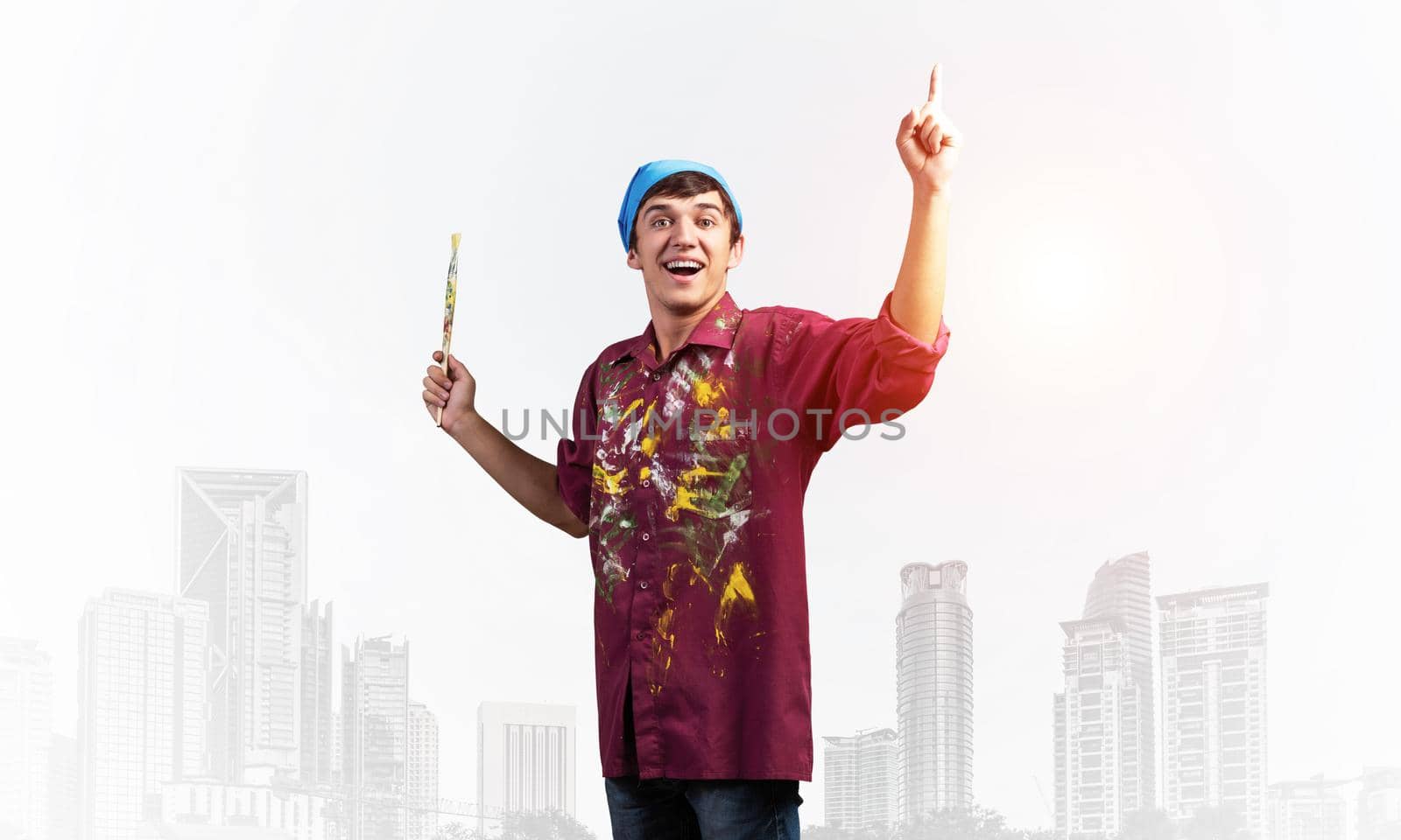 Young emotional artist gesturing with paintbrush. Happy painter in shirt and bandana standing on background modern office buildings. Creative hobby and artistic occupation concept with copy space