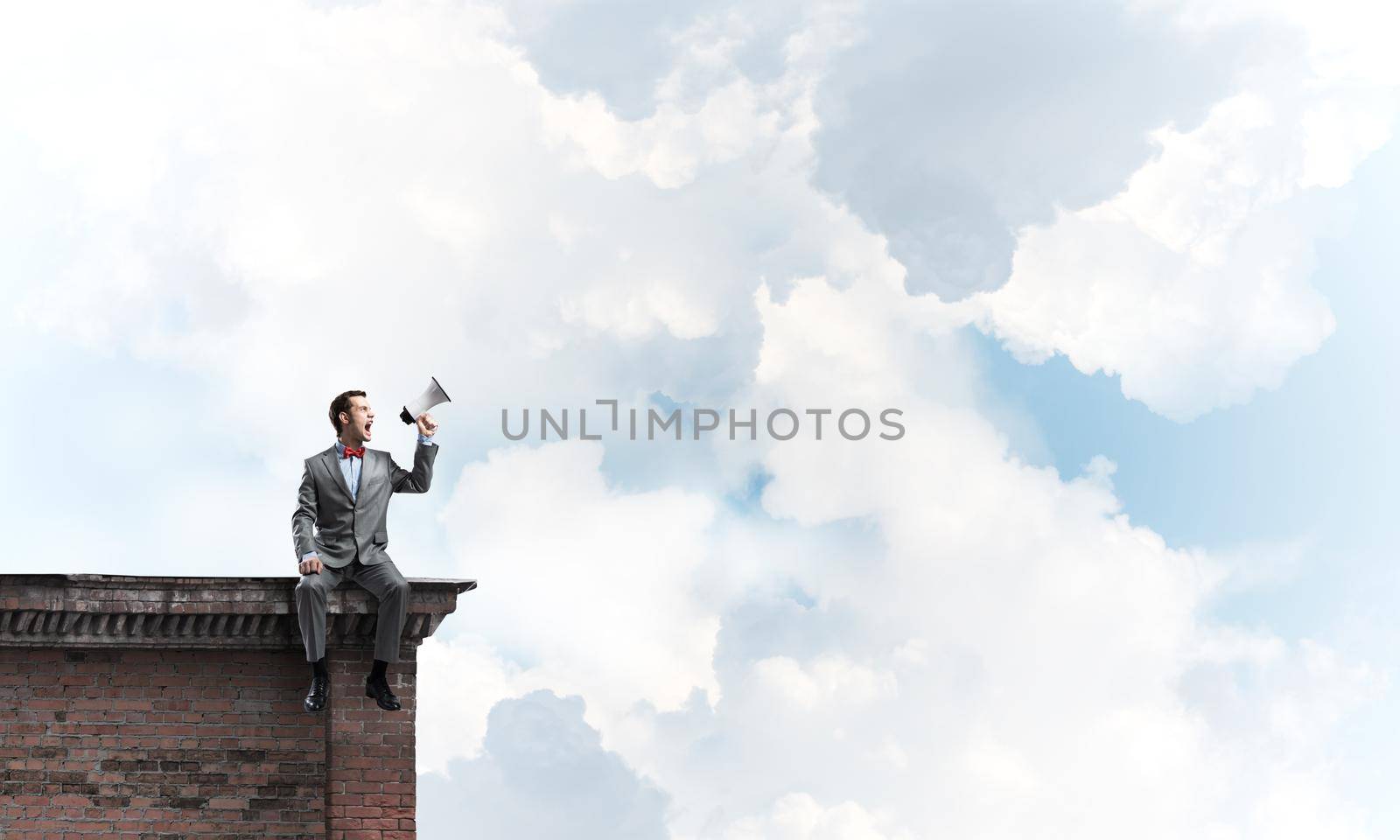 Young businessman sitting on roof and screaming emotionally in megaphone