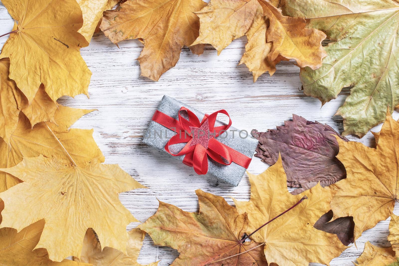Flat lay cute composition with gift box and yellow autumn leaves. Holiday present decorated red ribbon bow on vintage wooden desk. Happy thanksgiving congratulation. Still life of autumn season.