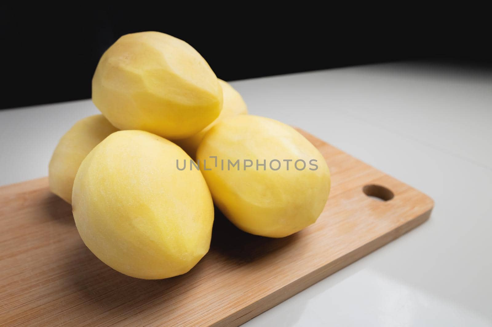 Some peeled potatoes on a wooden cutting board. On a white table against a dark background.