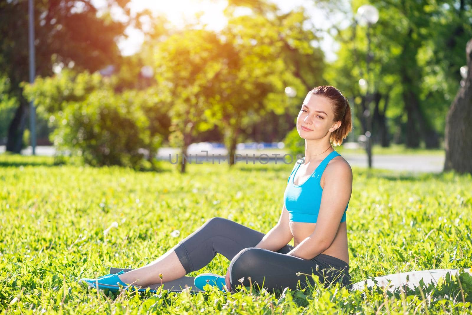 Beautiful smiling girl relax in park at sunrise. Portrait of young charming woman sitting on green grass. Training and meditation outdoor. Morning daily exercises. Happiness and harmony with nature