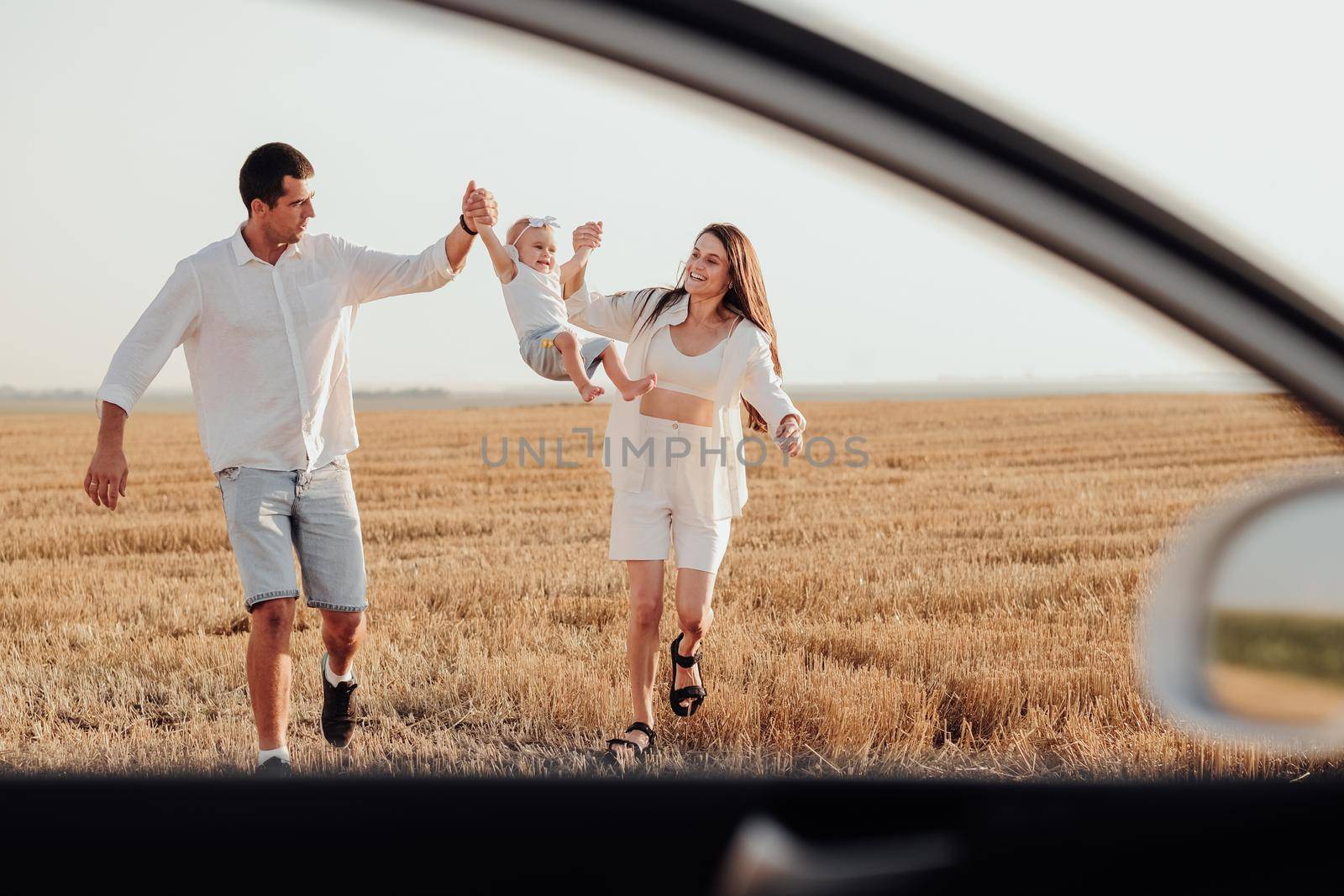 View Through Car Window, Mom and Dad Holding Their Toddler Daughter by Hands While Walking on Field at Sunset