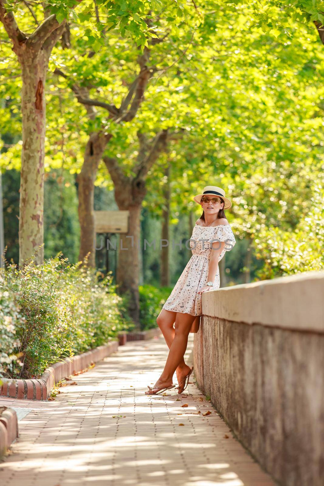 The bright beautiful girl in a light dress and hat walks along alley of trees in Monaco in sunny weather in the summer, handbag in hand. High quality photo