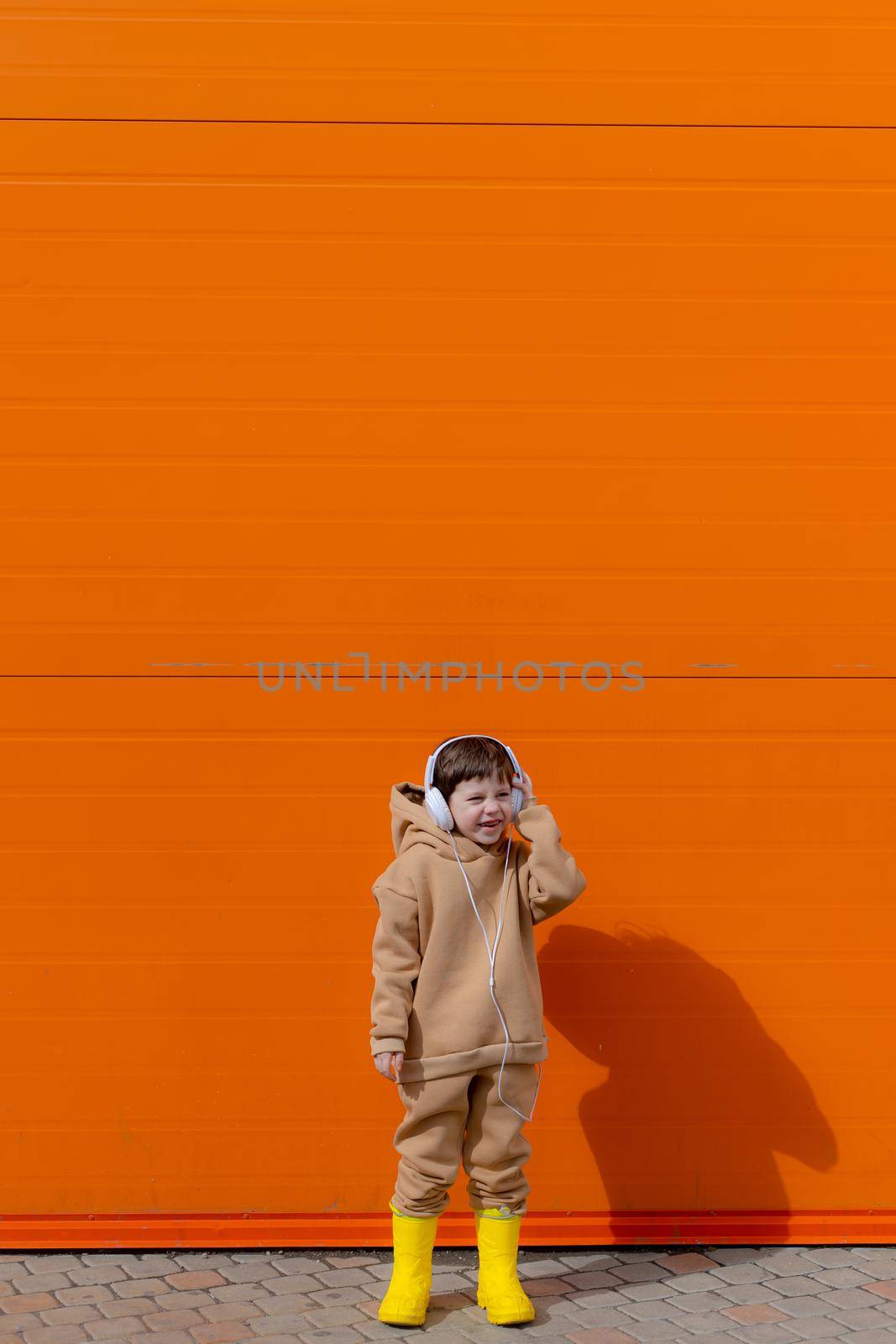 A boy listens to music with headphones on the background of an orange wall . Copy space. Mockup. An article about modern children. An article about modern music. Article about the choice of headphones. Urban lifestyle