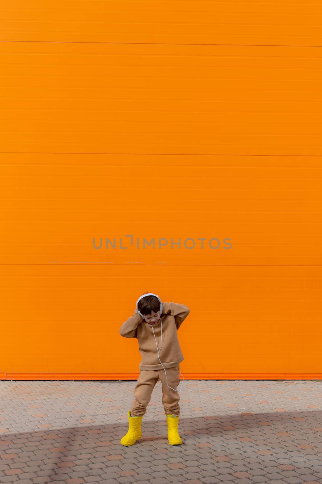 A boy listens to music with headphones on the background of an orange wall . Copy space. Mockup. An article about modern children. An article about modern music. Article about the choice of headphones. Urban lifestyle