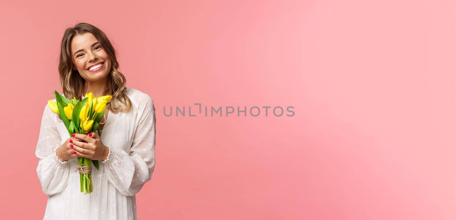 Holidays, beauty and spring concept. Portrait of lovely caucasian blond girl in white dress, smiling upbeat, holding yellow tulips, having perfect romantic date with flowers as gift, pink background.