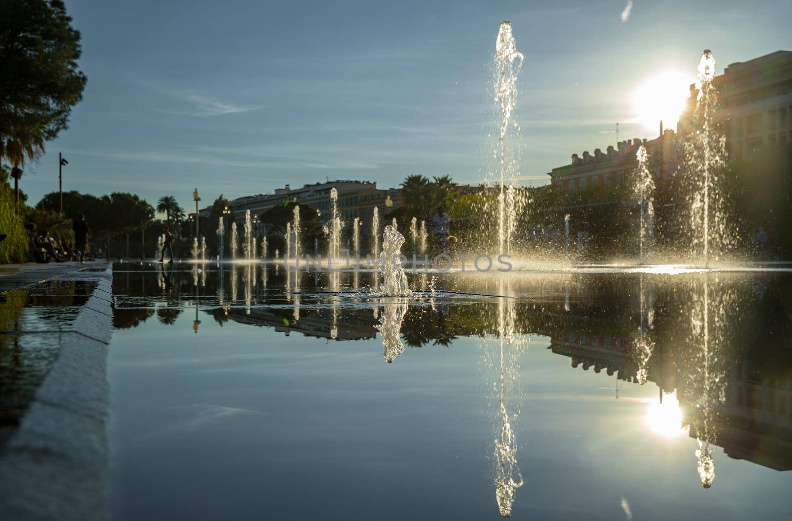 France, Nice, Famous place of Nice, square of fountain, mirror of water, playing kids, walking couples, a couple of old people are basking in the sunset, sun reflection, passing tram. High quality photo