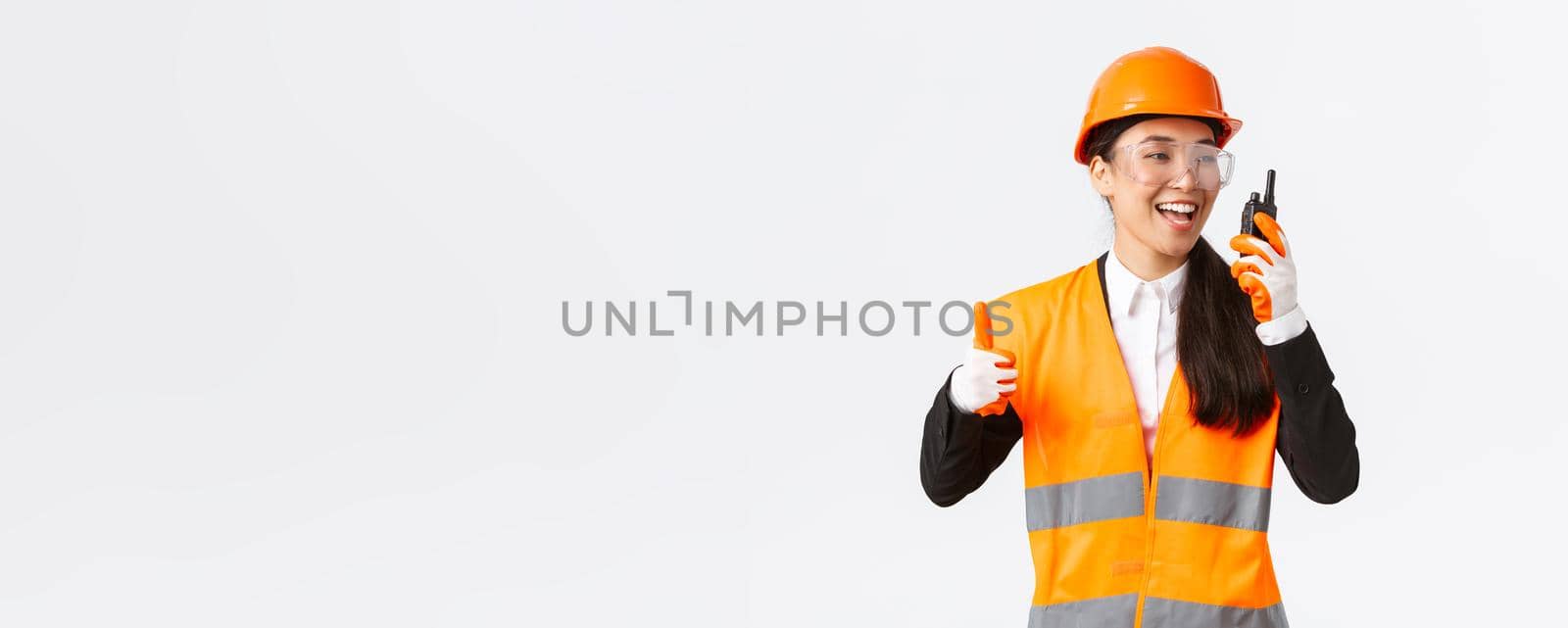Satisfied happy smiling asian female engineer, industrial technician in safety helmet and uniform showing thumbs-up while praising great work using walkie-talkie, give permission to work.