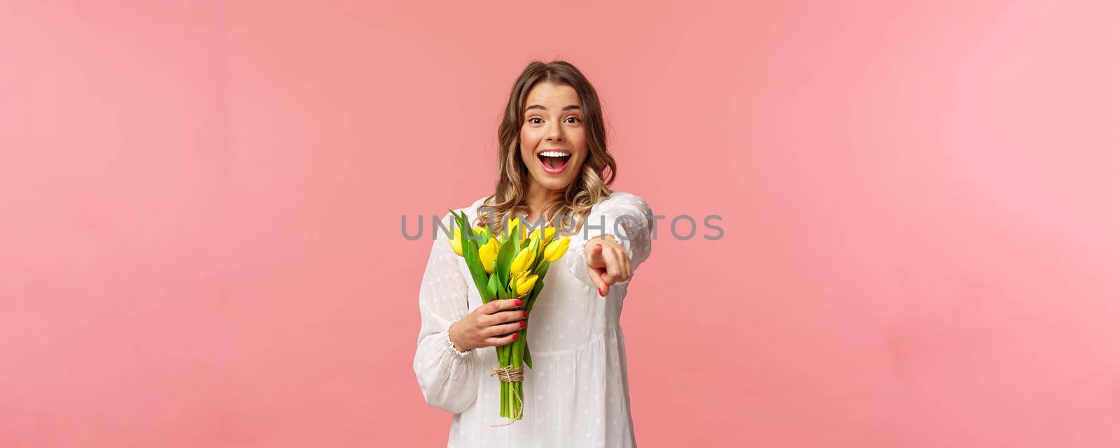 Holidays, beauty and spring concept. Portrait of excited happy and upbeat young blond girl in white dress, holding bouquet of yellow tulips, pointing finger at camera amazed, pink background by Benzoix