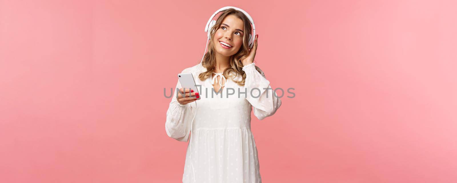 Portrait of tender, beautiful blond caucasian girl in white dress, daydreaming while listening favorite romantic track, wear headphones, hold mobile phone look up thoughtful, enjoy favorite track.