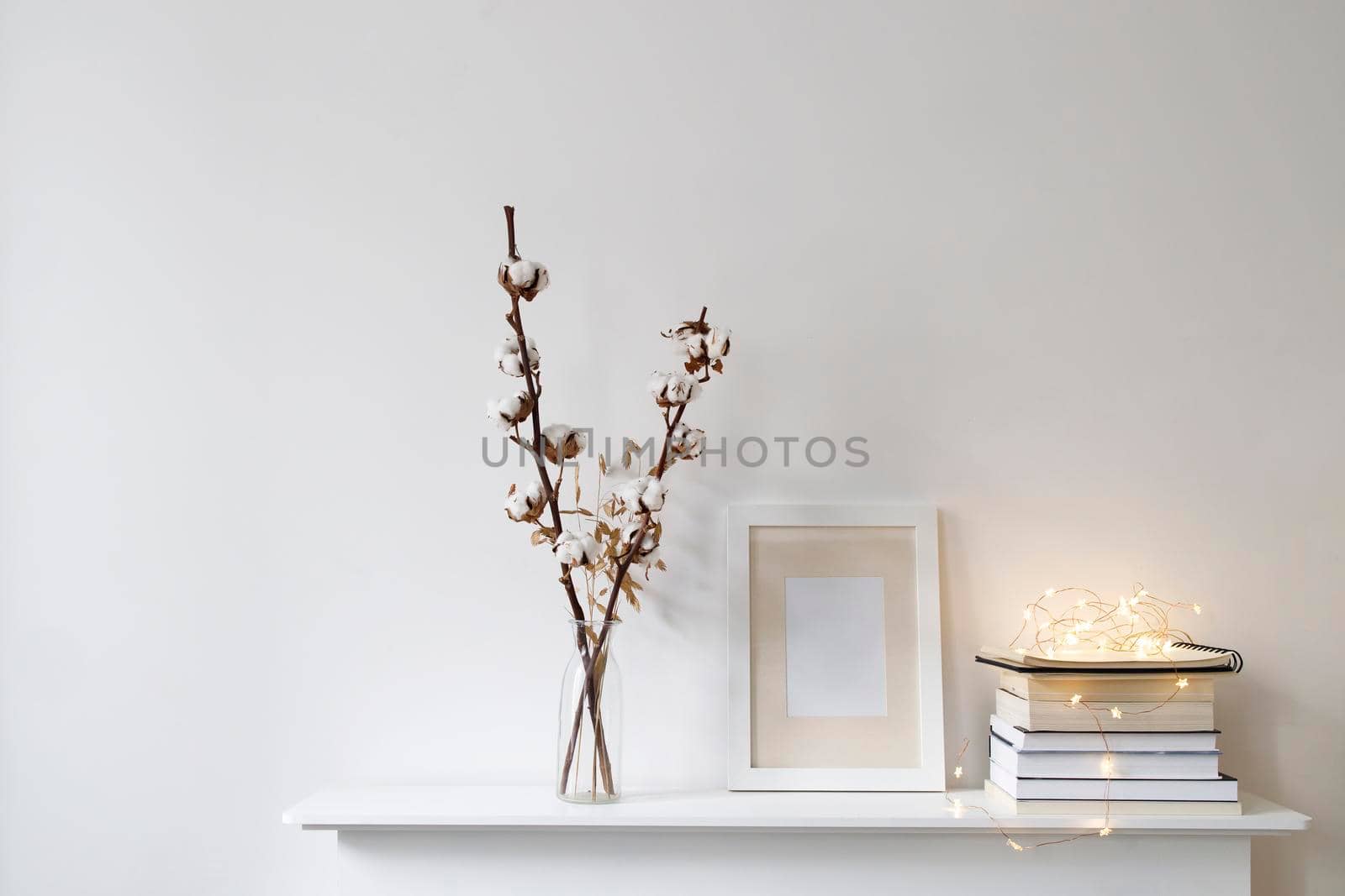 Scandinavian style room interior in white tones. A vase with cotton flowers, a stack of books, a photo frame, a garland on a wooden surface of a shelf. Copy space.